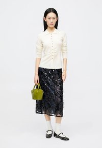Cream knitted cardigan with button front, paired with a black sequined skirt. Model holds a small green handbag and wears white socks and black shoes.
