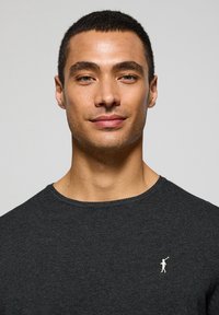 Jersey de cuello redondo en gris oscuro con un tejido texturizado, con un pequeño logo blanco en el lado izquierdo del pecho. Tela suave, diseño ajustado.