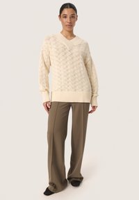 Crème gebreide trui met ruitpatroon, geribde halslijn en manchetten. Gepaard met losse beige broek en donkere schoenen, staat het model tegen een neutrale achtergrond.