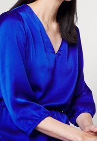 Blusa de satén azul con escote en V, mangas abullonadas de tres cuartos y cintura ajustada. Textura suave con un ligero brillo.