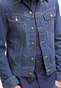 Giubbotto in denim blu con un design classico, chiusura con bottoni, due tasche al petto e una texture leggermente sbiadita. Accentuato da accessori in oro.