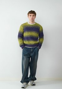 Chunky gebreide trui in groen en paarse strepen, gecombineerd met losse blauwe denim jeans en grijze sportschoenen. Minimalistisch ontwerp met textuurstof.