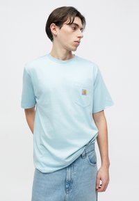 Νεαρός άνδρας με ανοιχτό μπλε μπλουζάκι Carhartt και τζιν τζιν, κοιτάζοντας προς τα αριστερά του, με φόντο λευκό κενό.