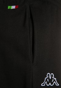 Czarne dresowe spodnie z bocznymi kieszeniami, z haftowaną włoską flagą i logo Kappa w białym i niebieskim kolorze. Miękka faktura, dopasowany krój.