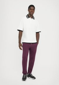 Camisa polo branca com detalhes pretos e uma gola gráfica, combinada com calças de moletom roxas e sapatilhas de desporto pretas.