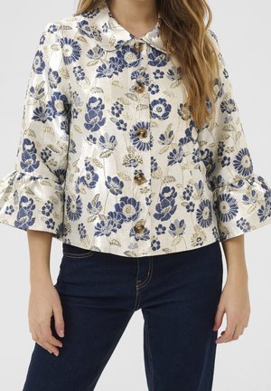 Femme portant une veste crème avec un motif floral bleu et doré, manches trois-quarts volantées et boutons marron, associée à un jean bleu foncé.