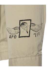 Beige stof med et trykt design af en faldskærm og vinger, med teksten "APO" og "710" i sort. Mat tekstur, ingen yderligere udsmykninger.