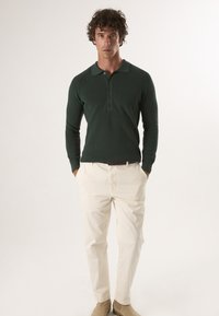 Dunkelgrünes Langarm-Poloshirt mit gerippter Textur, Knopfleiste und leichten weißen Hosen, getragen mit hellbeigen Schuhen.