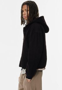Sort zip-hoodie med hætte, lavet af blødt stof. Har forlommer og en afslappet pasform, lagdelt over en ternet skjorte.