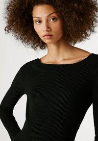 Juodas raukšlėtas megztinis su plačiu iškirpimu ir ilgomis rankovėmis. Audinys turi švelnų blizgesį, pabrėžiantį jo tekstūrinį raštą.