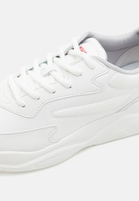 White sneaker with textured synthetic upper, smooth rubber sole, padded collar, and grey accents. Features white laces and logo detail.