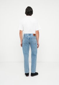 Világoskék farmernadrág, egyenes fazonban, barna bőr folttal és minimális varrási részletekkel. Fehér pólóval viselve.