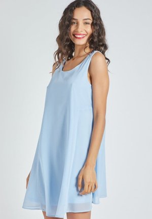 Mujer joven con cabello rizado sonriendo, vistiendo un vestido azul claro sin mangas hasta la rodilla, de pie contra un fondo claro y liso.