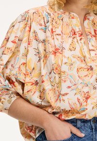 Haut floral avec un fond crème, présentant des motifs vibrants orange, jaune et bleu. Coupe ample, tissu texturé et un col boutonné.
