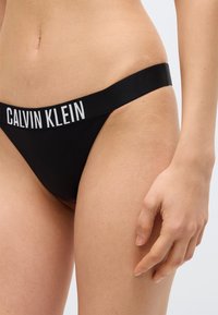 Γυναίκα που φοράει μαύρα εσώρουχα Calvin Klein με λογότυπο στη μέση, εμφανίζοντας τη μέση, το ισχίο και το δεξί χέρι της σε λευκό φόντο.