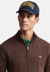 Bruine kabelgebreide trui met een ritssluiting aan de kraag, gedragen over een groen poloshirt. De marineblauwe pet heeft gele letters met "GANT 1949".