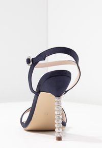 Námořnické saténové sandály na podpatku s kotníkovým páskem, zdobené dekorativním podpatkem posetým krystaly a hladkým, matným povrchem.