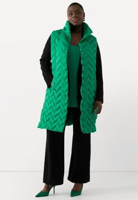 Grön quiltad lång väst med hög krage, försedd med tryckknappar. Kombination med en grön topp och svarta vida byxor, fulländad med gröna klackar.