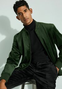 Giacca bomber verde in velluto con un dolcevita nero e pantaloni neri dalle texture, seduto contro uno sfondo chiaro. Leggera lucentezza e vestibilità aderente.