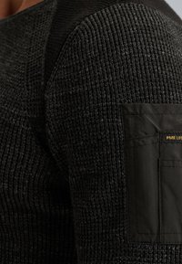 Close-up van een zwart getextureerde gebreide mouw van een trui met een vastgenaaide zwart stoffen patch zak, voorzien van een klein geel "PME LEG" label.