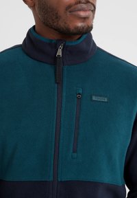 Blue and navy fleece jacket with a high collar, zip closure, and chest pocket. Features a TAG24 logo and soft, textured fabric.