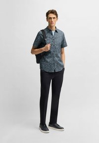 Camicia a maniche corte blu navy con un motivo floreale, abbinata a pantaloni neri e sneakers scure; uomo che tiene una borsa a tracolla nera.