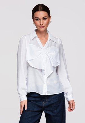 Kvinde iført en hvid bluse med lange ærmer og en stor sløjfe, samt mørkeblå jeans, stående foran en ensfarvet baggrund.