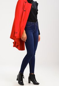 Czerwony trencz, czarna strukturalna bluzka, niebieskie dopasowane dżinsy i czarne botki za kostkę z przodem na sznurówki.