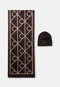Black and beige scarf with geometric pattern next to matching black beanie hat with a square embroidered logo patch.