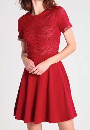 Femme portant une robe rouge avec un corsage ajusté à motif de dentelle, des manches courtes et une jupe évasée, debout devant un fond uni.