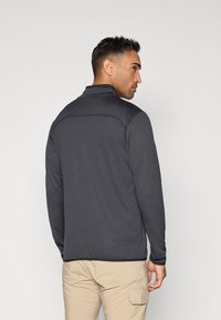 Meeste tumehall zip-up jakk tekstuuriga mustriga, kõrge krae ja pikad varrukad, koos heledate beeži pükstega.