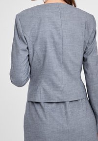 Grå skräddarsydd blazer med en texturerad yta, med fyrkantig halsringning och strukturerad rygg, designad för professionellt bruk.