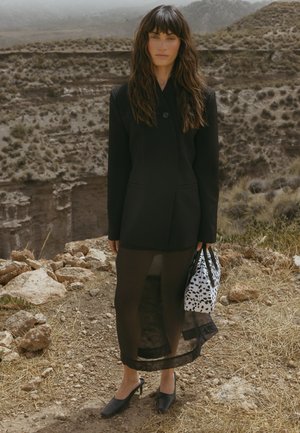 Blazer negro ajustado con un solo botón, superpuesto a una falda negra translúcida y combinado con tacones negros y un bolso con estampado.