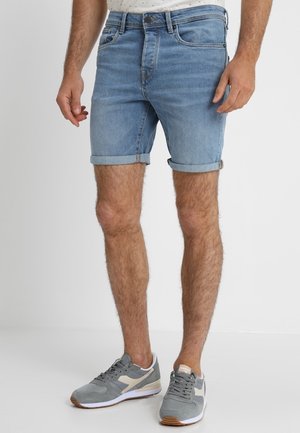 Homme portant un short en jean bleu clair roulé et des baskets grises, debout sur un fond blanc uni.