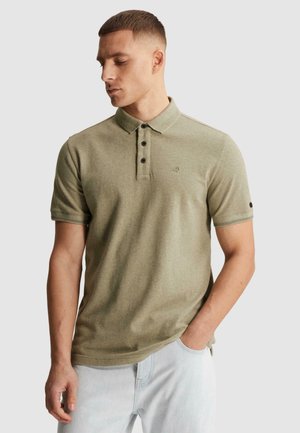 Jeune homme aux cheveux courts portant un polo à manches courtes vert olive clair et un jean bleu clair, regardant vers la gauche avec une main dans la poche.