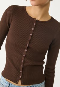 Hnědý žebrovaný pletený cardigan s kulatým výstřihem, dlouhými rukávy a osmi předními knoflíky v světlejším odstínu. Nabízí přiléhavý tvar a texturu.