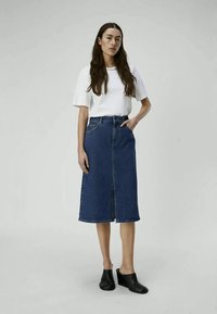 Saia de ganga com uma costura frontal e bolsos laterais, combinada com uma t-shirt branca. A modelo usa mules pretos de salto, exibindo um conjunto casual.