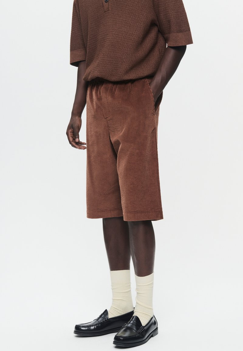 Shorts en velours côtelé marron avec une coupe décontractée, dotés de poches latérales, associés à des chaussettes côtelées de couleur claire et des mocassins noirs.