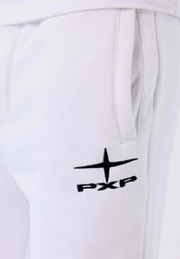 Primo piano di una tasca di pantaloni sudati bianchi con il logo PXP nero e un emblema a stella ricamato sotto l'apertura della tasca.