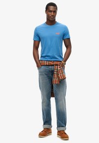 T-shirt di cotone blu con logo ricamato, abbinata a jeans in denim chiaro arrotolati. Una camicia a quadri legata in vita e scarpe marroni aggiungono dettagli.