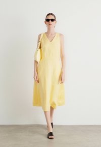Rochie galbenă din in, cu decolteu în V și siluetă în A. Modelul poartă o geantă mică albă și sandale negre cu talpă plată, precum și ochelari de soare.