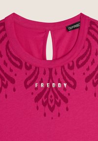 Camiseta fucsia con cuello redondo y apertura en la parte posterior, que presenta un patrón decorativo tonal y el texto plateado "FREDDY" en la parte delantera.