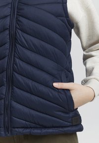 Tmavě modrá prošívaná vesta na zip, s diagonálním prošíváním, bočními kapsami a značkovou štítkem na lemu. Měkký, lehký materiál.