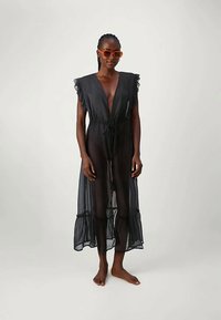 Γυναίκα φορώντας ένα διαφανές μαύρο φόρεμα με φούσκες στα μανίκια και το στρίφωμα, βαθύ V-λαιμόκοψη, κ tied στη μέση, και πορτοκαλί γυαλιά ηλίου, στέκεται ξυπόλυτη.