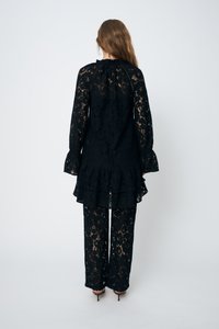 Completo in pizzo nero composto da una lunga tunica con orlo arricciato e maniche a campana, abbinato a pantaloni palazzo coordinati.