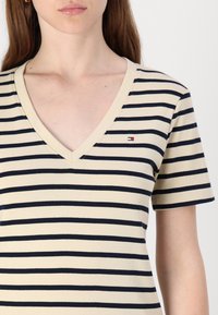 Beige V-hals T-skjorte med marineblå horisontale striper. Har korte ermer og et lite logo-merk på venstre bryst. Mykt bomullsmateriale.