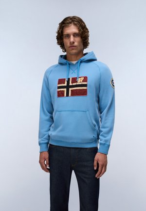 Young man with curly hair wearing a light blue hoodie with red, white, and black flag patch and dark jeans, standing against a plain background.