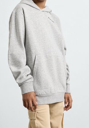 Person iført en lysegrå hættetrøje med lomme foran og beige cargobukser, stående mod en ensfarvet lys baggrund.