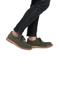 Zapatos de ante verde con detalles en cuero marrón, diseño de cordones y costuras blancas. La suela de goma tiene textura para un mejor agarre. Se llevan con jeans oscuros.
