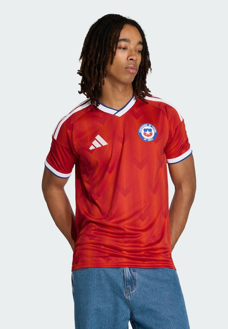 Młody mężczyzna w czerwonej koszulce piłkarskiej Chile z logo Adidas, stoi z rękami za plecami, patrząc w dół na bok.